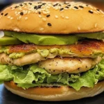 A close-up of a juicy Avocado Chicken Burger on a toasted brioche bun. The grilled chicken breast is golden-brown with slight charring, topped with creamy mashed avocado, fresh lettuce, ripe tomato slices, and thinly sliced red onions. A drizzle of garlic aioli adds a rich finish. Served on a rustic plate with a side of crispy fries. Warm, natural lighting highlights the textures and colors for an appetizing, homemade feel.